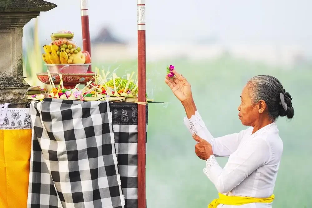 An elderly Balinese woman in traditional white ceremonial dress praying with a flower in her hand in front of a temple shrine with fruit offerings, representing the living culture of Bali.