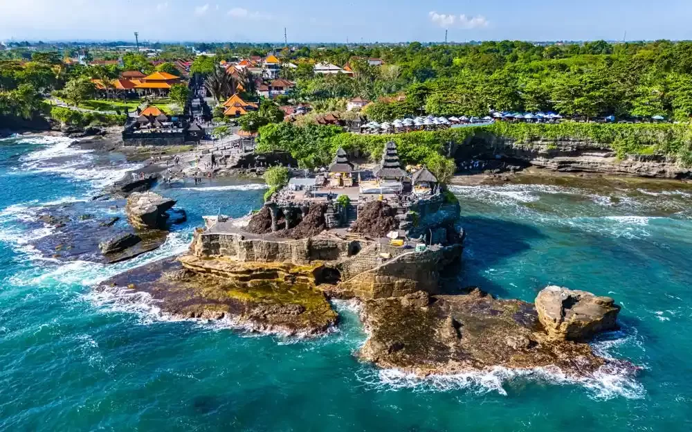 An aerial view of the ancient Tanah Lot temple complex in Bali, perched on a rock formation in the sea with waves crashing around it, and the vibrant coastline and village in the background.