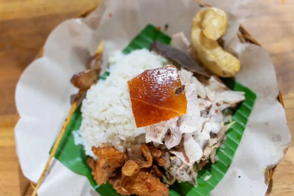 A close-up top-down view of a babi guling dish, a classic Bali street food, served on a banana leaf. The plate includes a mound of white rice topped with crispy pork skin and shredded suckling pig.