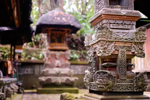 A close-up of an ornate, moss-covered stone carving at a Balinese temple, a key element of traditional Balinese architecture. The background shows another traditional shrine in a lush setting.