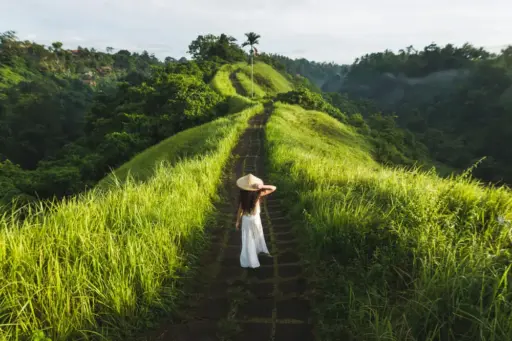 A woman in a white dress and straw hat walks along the stone path of the Campuhan Ridge Walk in Ubud, Bali, surrounded by lush green grass and jungle valleys.