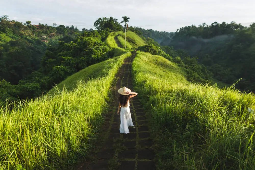 A woman in a white dress and straw hat walks along the stone path of the Campuhan Ridge Walk in Ubud, Bali, surrounded by lush green grass and jungle valleys.