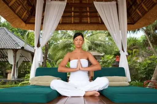 A woman in white meditating in a yoga pose on a daybed inside a traditional Balinese bale, representing a mindful Bali wellness and slow travel experience.