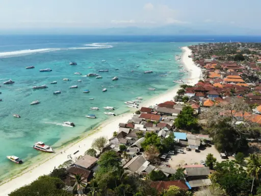 Aerial view of Jungut Batu beach on Nusa Lembongan, showing the long white sand beach, turquoise water filled with tour boats, and the coastal village. The Bali mainland is visible on the horizon.