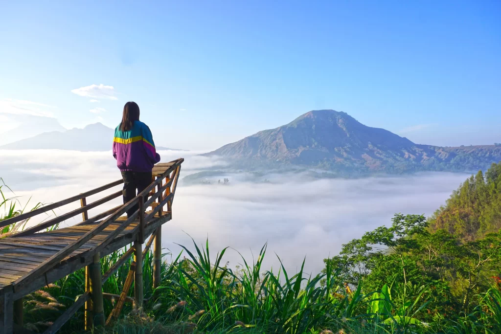 A woman stands on a wooden viewing platform, looking out over a sea of clouds covering the valley with the large Mount Batur volcano rising in the distance under a clear sky.