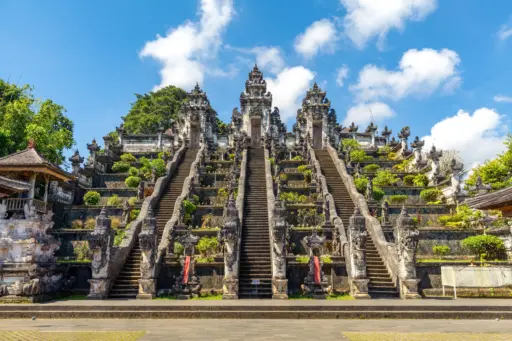 A grand, symmetrical view of Besakih Temple with a long stone staircase leading up to tiered shrines and ornate statues, surrounded by colorful flowers and a blue sky with clouds.