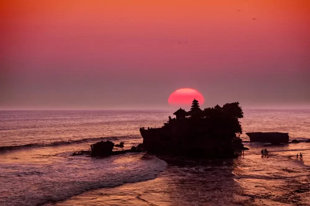 The Tanah Lot temple in Bali silhouetted against a dramatic sunset, with the large red sun setting directly behind the temple's tiered roofs over the ocean.