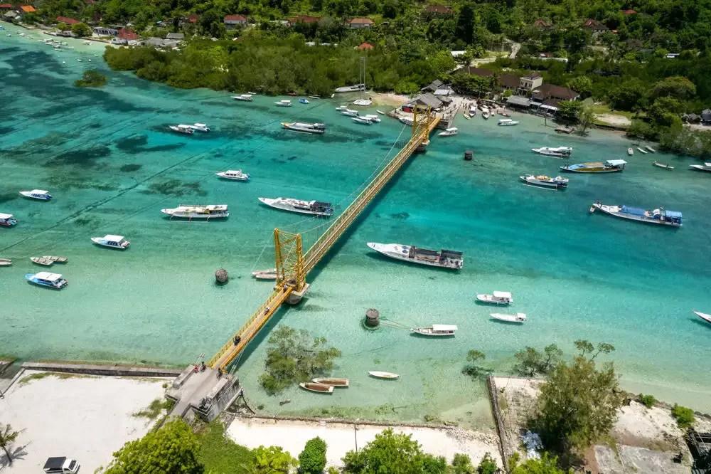 An aerial view of the iconic Yellow Bridge connecting Nusa Lembongan and Nusa Ceningan, with dozens of boats moored in the clear turquoise water below.