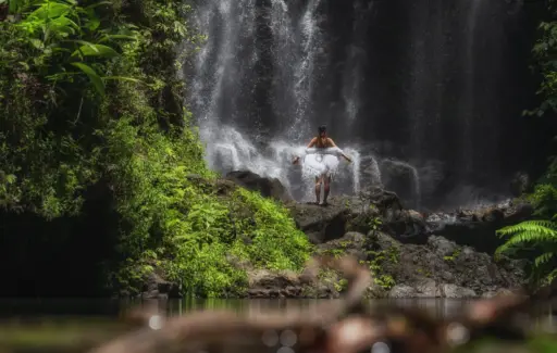 A woman in a white dress stands on a rock with her arms out, facing a powerful waterfall in a lush, green jungle setting.