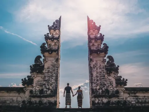 A couple holding hands, silhouetted against a bright sky, stands between the ornate "Gates of Heaven" at Lempuyang Temple in Bali