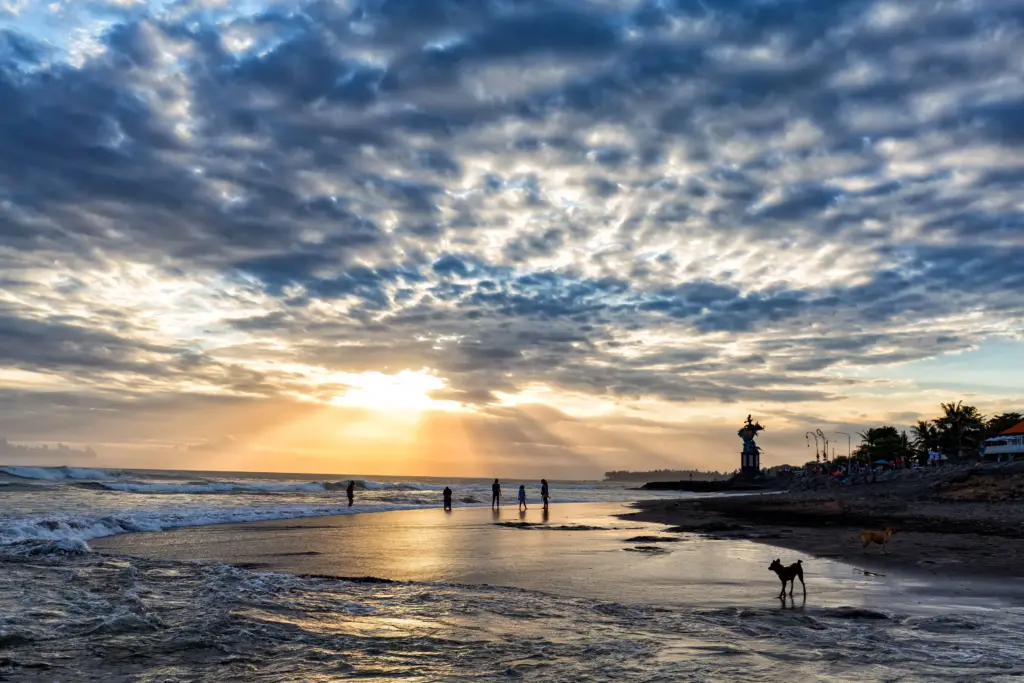 A beautiful sunset over the ocean at Pererenan Beach, with sunbeams breaking through the clouds, silhouetted people walking on the wet sand, and two dogs playing nearby.