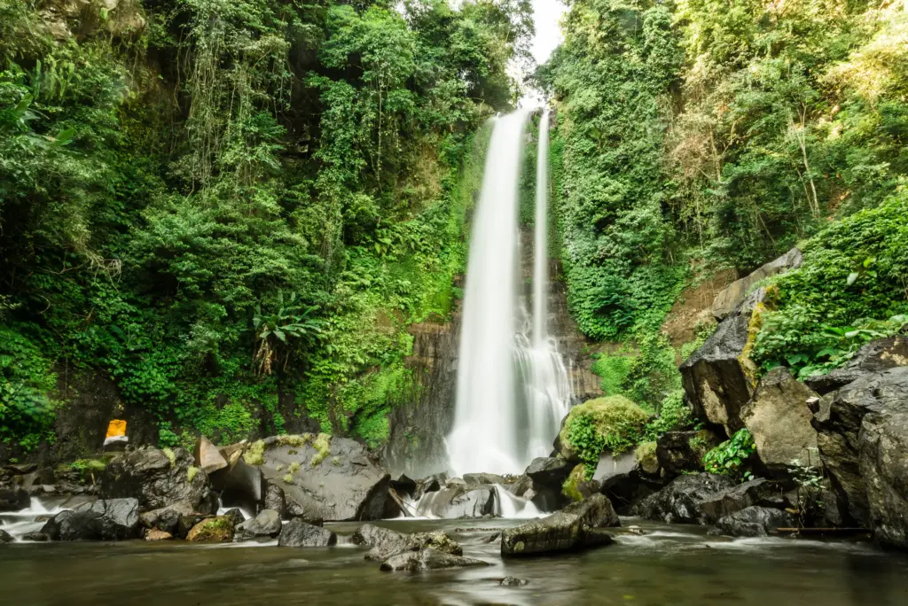 A long-exposure shot of a powerful waterfall cascading down a rock face surrounded by dense, lush green jungle and large mossy rocks in a river at its base.