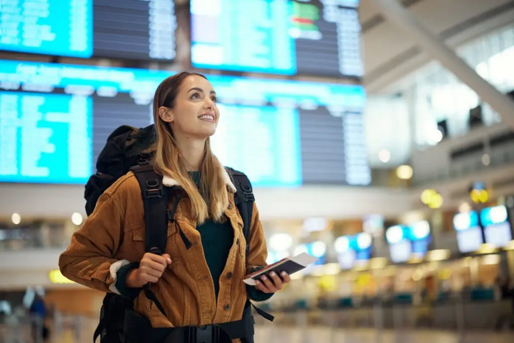A smiling young woman with a backpack, holding a passport and a boarding pass in a bright airport terminal, looking up at a flight information screen.