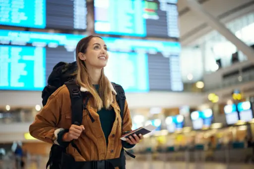 A smiling young woman with a backpack, holding a passport and a boarding pass in a bright airport terminal, looking up at a flight information screen.