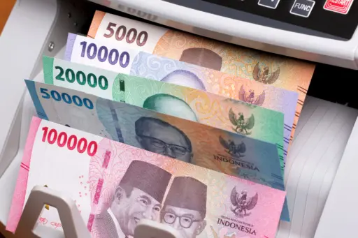 A close-up of a stack of Indonesian Rupiah banknotes in various denominations (100,000, 50,000, 20,000, 10,000, and 5,000) being counted by a money counting machine.