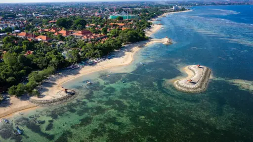 An aerial shot of the calm, clear turquoise waters and soft golden sand of Sanur Beach, showing small breakwaters and resorts nestled among green trees.